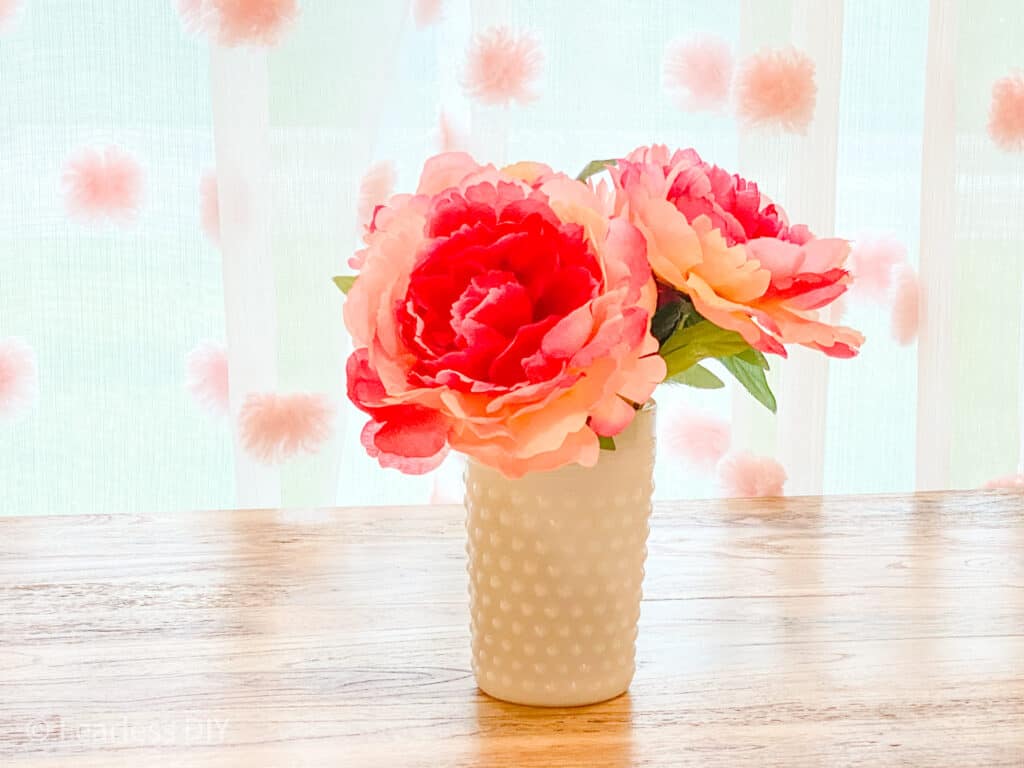 pink peonies in a white hobnail jar on a wooden childs table in front of shear curtains with pink puffballs staged by fearlessdiy.com
