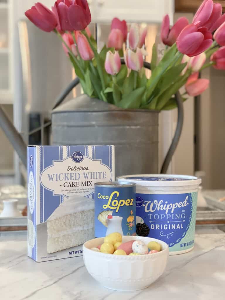 Boxed cake mix, cream of coconut, tub of whipped topping and Robin's Egg candy in front of a galvanized watering can filled with pink tulips