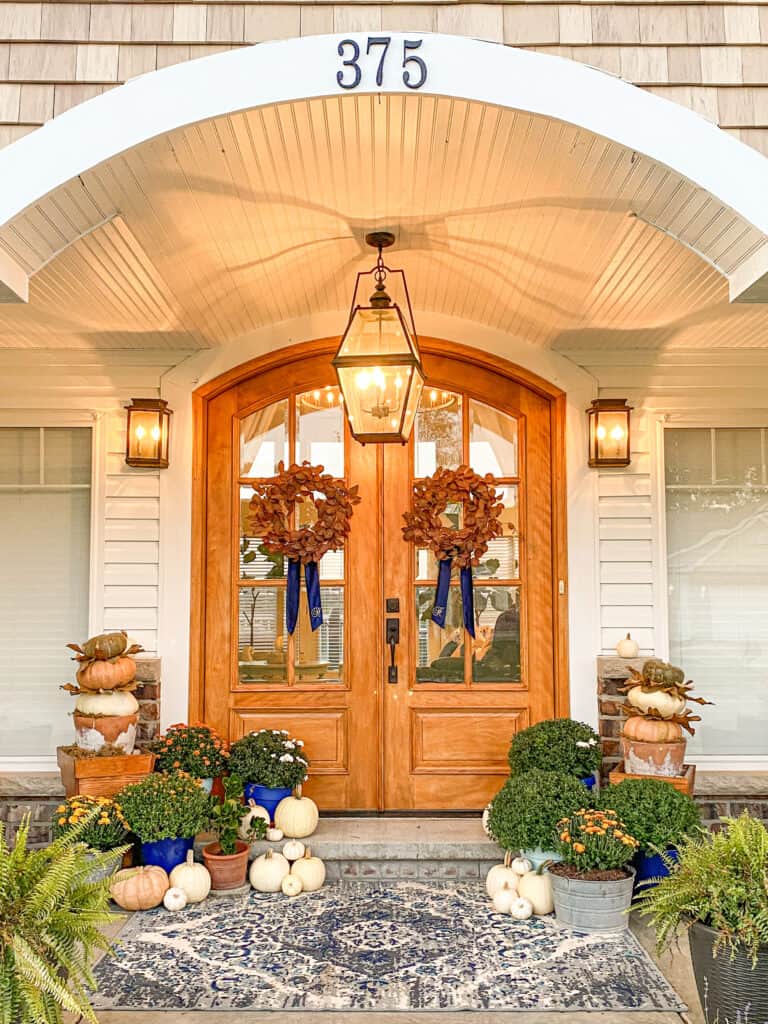 arched wooden double front doors with fall wreaths with blue velvet wreath sashes. The door is framed by mums, white, orange and green pumpkins.