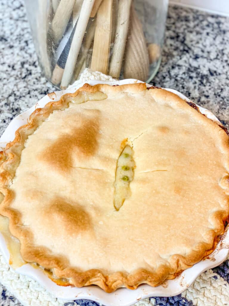 Flaky crust with chicken pot pie oozing out the center in a white stoneware pie plate in front of a jar with wooden spoons 
