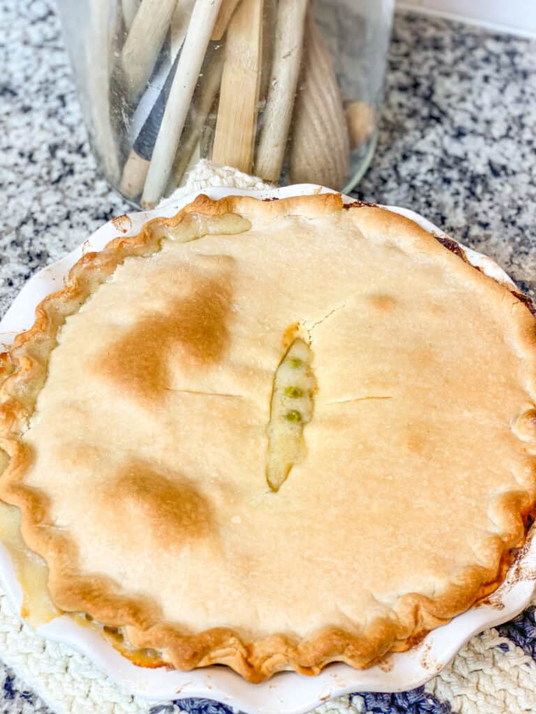 Flaky crust with chicken pot pie oozing out the center in a white stoneware pie plate in front of a jar with wooden spoons