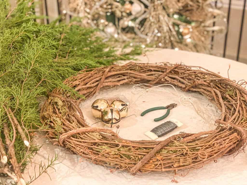 grapevine garland, floral wire, wire cutters, battery operated lights, 3 3 inch bells and fresh evergreen clippings on a table in front of a sparkly Christmas tree. 