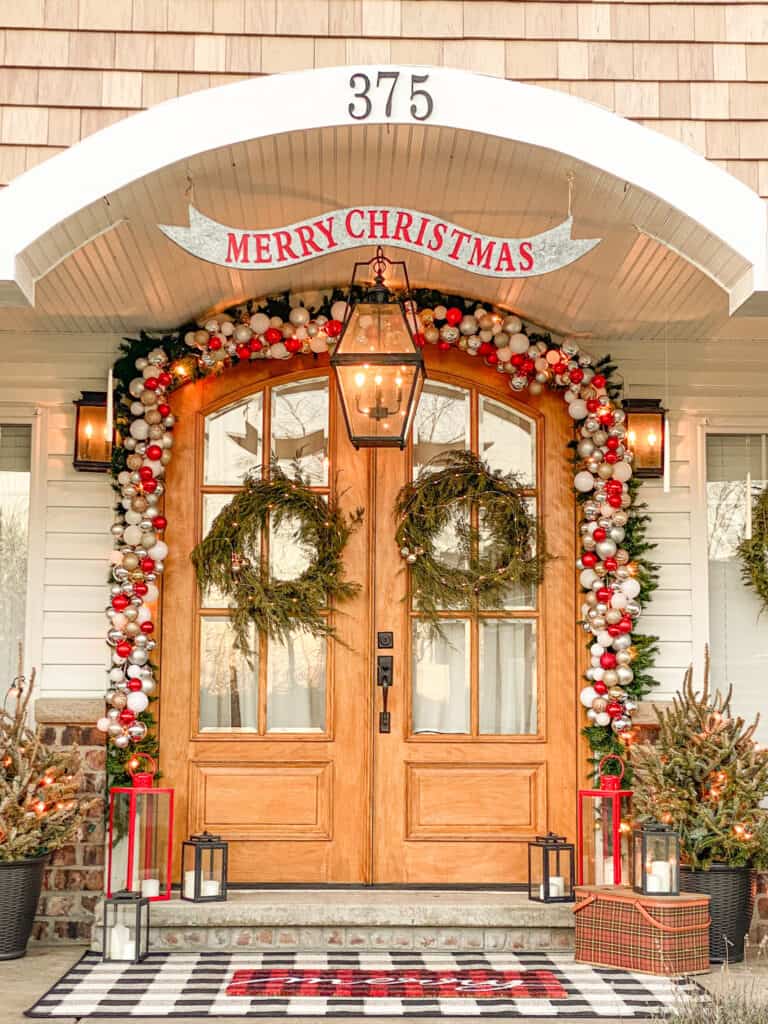 red, white, gold and silver ornament garland surrounds wooden double arched front doors, flanked by red and black lanterns and potted douglas fir trees with large old fashioned lightbulbs